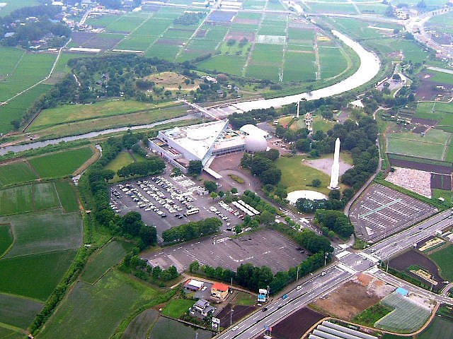 本館の右手前にH2ロケット、奥に風力発電装置、更に右奥に乗り物広場が見えます。川をはさんだ左に自然を生かした河川公園「風の広場」が隣接しています。